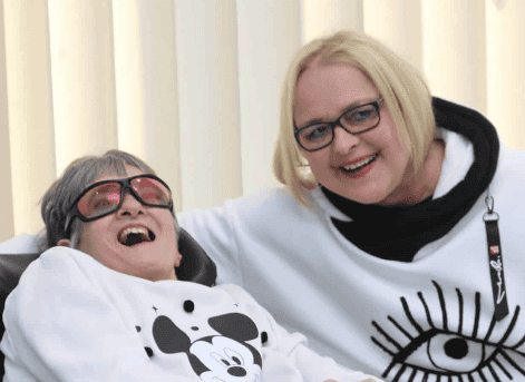Foto von einer lächelnden Frau mit Beeinträchtigung im Rollstuhl, neben ihr sitzt eine Mitarbeiterin der W f a A. Foto von einer lächelnden Frau mit Beeinträchtigung im Rollstuhl, neben ihr sitzt eine Mitarbeiterin der W f a A.